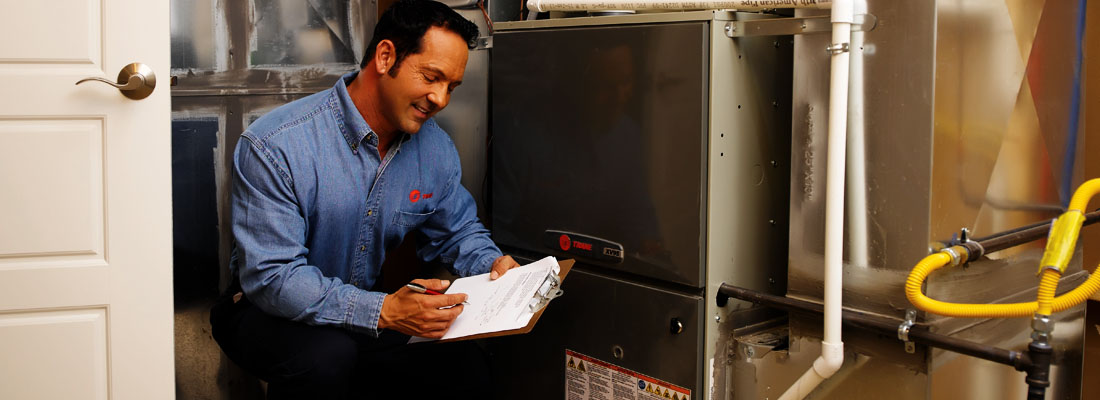 trane tech kneeling near furnace writing on clipboard